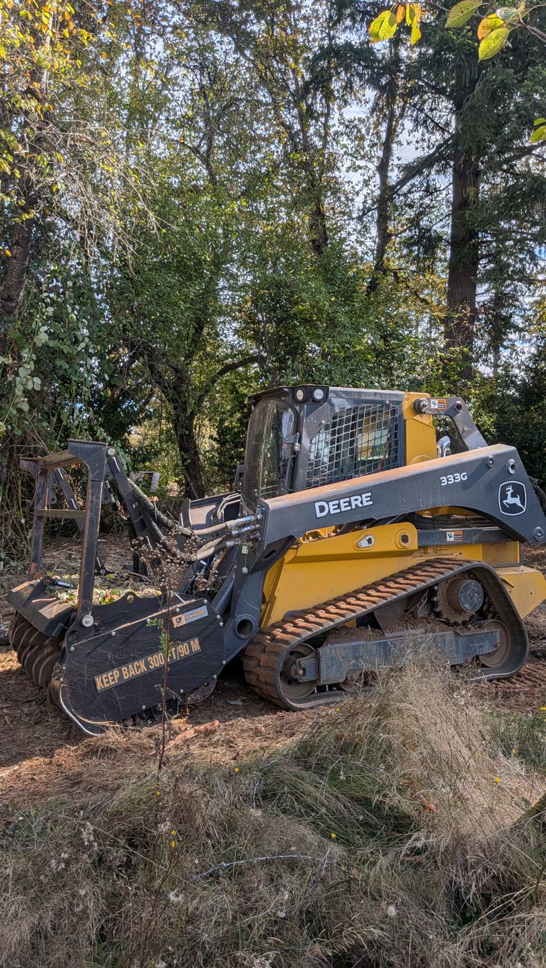 Close-up of a yellow John Deere 333G compact track loader with a forestry mulching head, standing among trees and brush